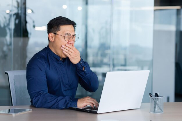 Een vermoeide jonge aziatische man met een bril zit op kantoor aan tafel en werkt aan een laptop hij