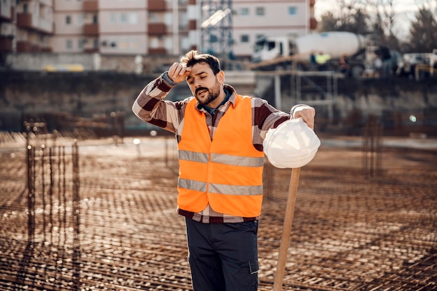 Een vermoeide werfmedewerker veegt het zweet af en rust uit van hard werken op de werf terwijl hij op de werf staat