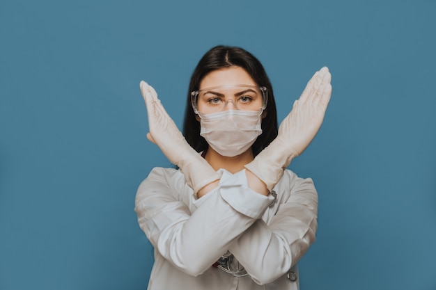 Een verpleegster met een beschermende bril, een chirurgisch masker en een medische jurk kruiste haar handen. Wat betekent dat we de pandemie van het coronavirus moeten stoppen.