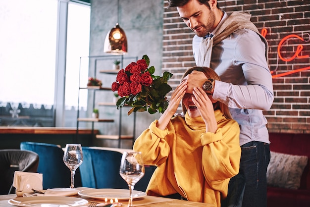 Een verrassing voor haar maken. man in blauw shirt verraste zijn mooie blonde vriendin in restaurant. hij houdt rode rozen bij zijn handen en sluit haar ogen. blonde jonge vrouw in mosterdtrui