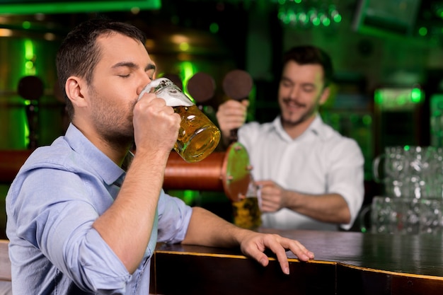 Een vers getapt biertje drinken. Knappe jonge man bier drinken terwijl barman bier tappen op de achtergrond