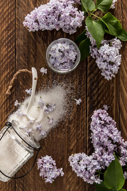 Een verticale weergave van zelfgemaakte suiker verspreid uit een glazen pot van lila bloemen een kom met bloemen lila takken houten bruine achtergrond