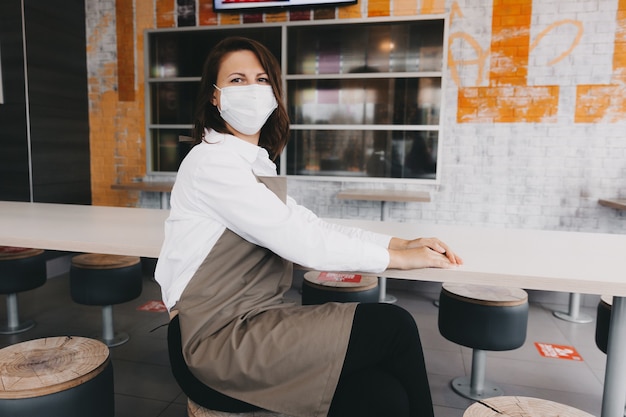 Een verveelde serveerster in een schort en een medisch masker zit in een café-bar. de eigenaar van de coffeeshop zonder bezoekers te bezoeken. bedrijf.