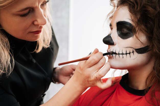 Een visagist verzint een demon voor een jong meisje in een schoonheidssalon. Halloween-vieringsconcept.