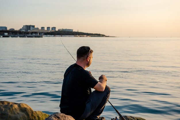 Een visser vangt vis in de zee met een hengel zittend op de kust van de rotsen bij zonsondergang