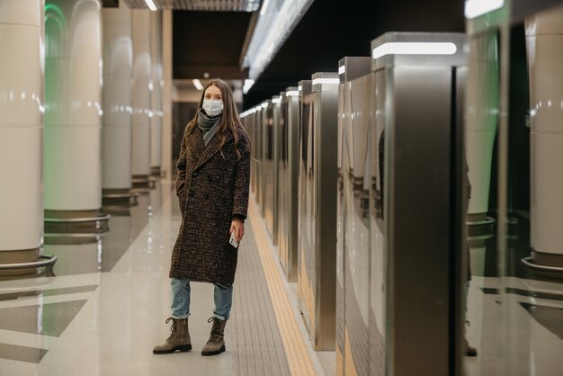 Een volledige foto van een dame met een medisch gezichtsmasker om de verspreiding van COVID te voorkomen, wacht op een trein en houdt een telefoon vast op het metroplatform. Meisje met chirurgisch masker houdt sociale afstand