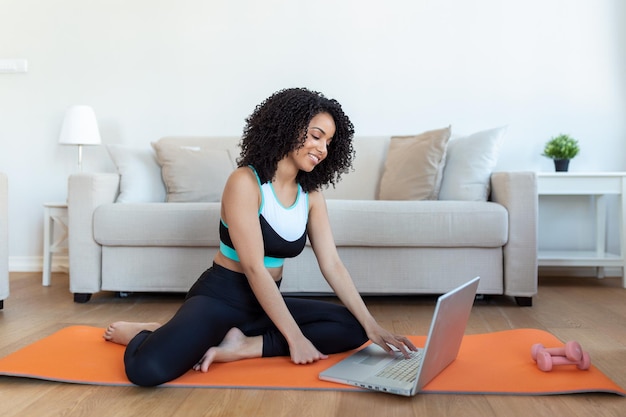 Een volwassen Afrikaanse vrouw doet yoga- en krachttrainingsoefeningen op een matje in haar woonkamer Ze volgt een online trainingsvideo op haar laptop