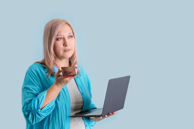 Een volwassen blonde vrouw van 3540 jaar oud communiceert met een smartphone en heeft een moderne laptop in haar handen die op een blauwe achtergrond staat