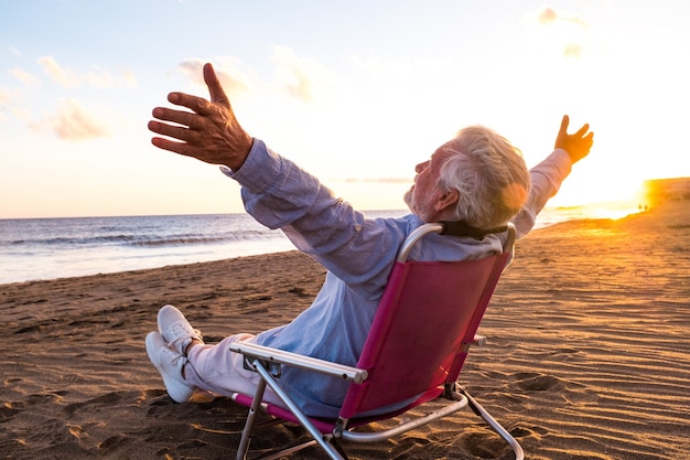 Foto een volwassen en oude man geniet van de zomervakanties alleen op het strand, zittend in een kleine stoel met uitzicht op de zee. mannelijke persoon die zich vrij voelt met geopende armen. vrijheidsconcept.
