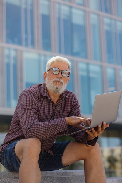 Een volwassen grijsharige man met een baard zit op kantoor en werkt op een laptop