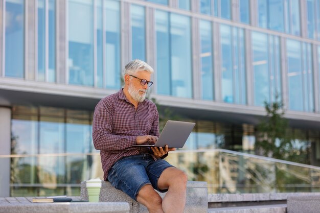 Een volwassen grijsharige man met een baard zit op kantoor en werkt op een laptop