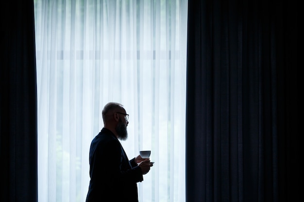 Een volwassen man met een baard drinkt koffie en kijkt uit het raam. Werkdag veld recreatie concept