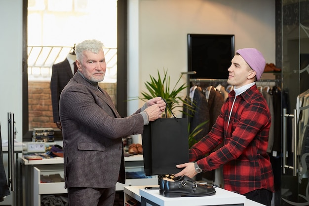 Foto een volwassen man met grijs haar en een sportieve lichaamsbouw neemt zwarte papieren zakken met aankopen van een verkoper in een kledingwinkel. een winkelbediende geeft papieren zakken aan een mannelijke klant.