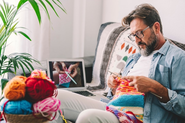 Een volwassen man thuis volgt online brei-tutorial om te ontspannen en te genieten van rust thuis vrijetijdsbesteding zittend op de bank - mannelijke mensen breien met computerklas hulp - mensen innerlijke levensstijl hobby