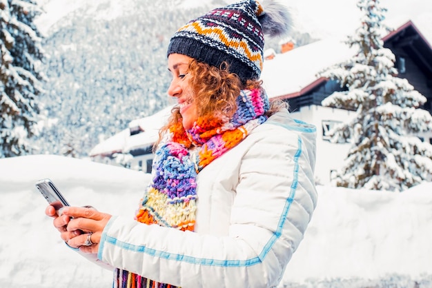 Eén volwassen vrouw die telefoon gebruikt in de winter sneeuw buiten vrije tijd moderne technologie activiteit alleen glimlachend en berichten wintervakantie vakantie seizoen vrouwelijke mensen buiten blauwe lucht en huis achtergrond