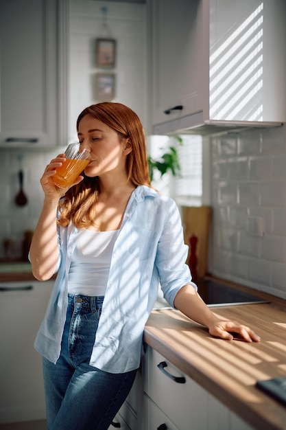 Een volwassen vrouw drinkt sinaasappelsap met haar ogen gesloten thuis.