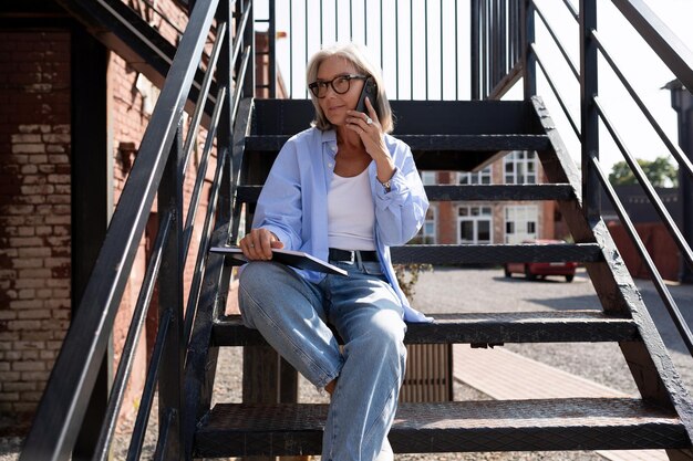 Foto een volwassen vrouw met grijs haar en bril controleert aantekeningen in een notitieboek terwijl ze op straat zit en