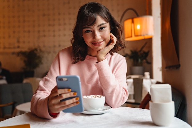 een vrij gelukkig lachende brunette jonge vrouw binnenshuis in café koffie drinken met behulp van mobiele telefoon.