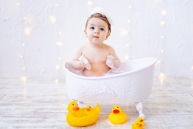 Een vrolijk babymeisje van zes maanden baadt in een bad met schuim en rubberen eenden, een klein kind heeft plezier bij het spelen met water het concept van zorg en hygiëne