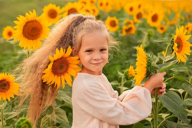 een vrolijk blond meisje in een roze linnen jurk staat in een veld met zonnebloemen