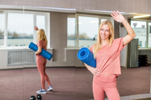 Een vrolijk blond meisje met een kleed opgerold in een rol zwaait met haar hand in een sportschool tijdens een training