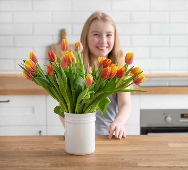 Een vrolijk en vrolijk meisje met een mooi boeket van rode en gele tulpen in een witte vaas op een houten aanrecht tegen een witte keuken achtergrond Lente tijd concept wazig beeld