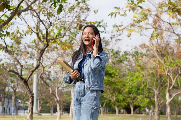 Een vrolijk jong meisje dat een mobiele telefoon gebruikt en een boek vasthoudt dat in het park staat, draagt een levensstijlconcept voor een jas en spijkerbroek