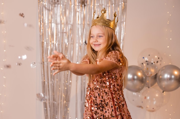 een vrolijk klein meisje in een elegante jurk en een kroon blaast confetti uit haar handpalmen en doet een wens.