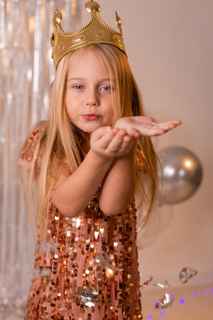 een vrolijk klein meisje in een elegante jurk en een kroon blaast confetti uit haar handpalmen en doet een wens.