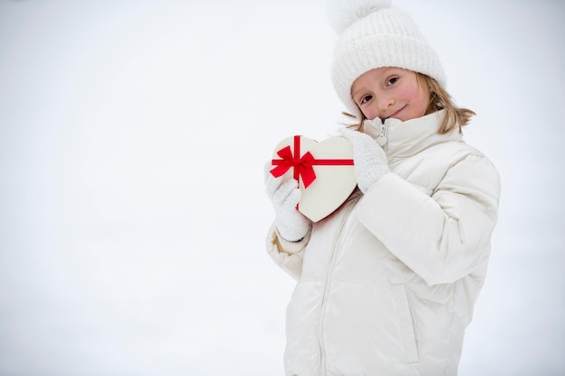 Een vrolijk klein meisje in witte winterkleren staat voor de sneeuw en houdt een hartvormige doos vast