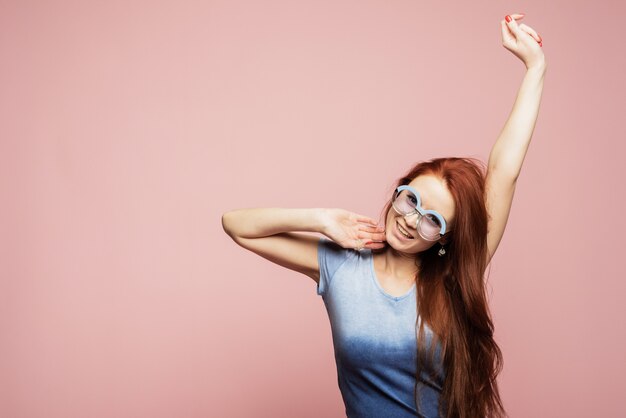 Een vrolijk meisje in blauwe ronde zonnebril verheugt zich en danst op een roze muur. Gelukkige mooie vrouw die op vakantie in de warme zomer gaat