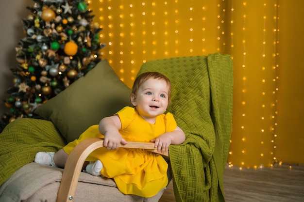 Een vrolijk meisje in een gele jurk zit in een schommelstoel met kerstboom