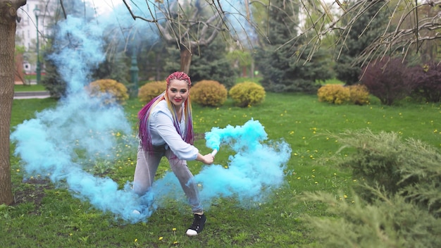 Een vrolijk meisje in een shirt en spijkerbroek met heldere regenboogvlechten en ongewone make-up. ze danst en verstopt zich in dikke blauwe kunstmatige rook tegen de achtergrond van het lentepark