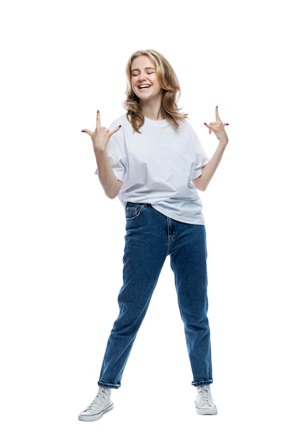Een vrolijk tienermeisje in spijkerbroek en een wit T-shirt staat en toont een geit met haar handen Energie en positiviteit Volledige hoogte Geïsoleerd op witte achtergrond Verticaal