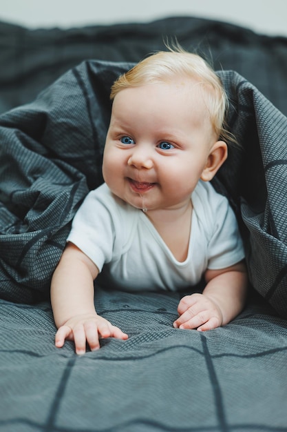 Een vrolijke baby jongen met mooie grote ogen ligt thuis op het bed in een wit bodysuit op grijs beddengoed het kind ligt thuis in het bed