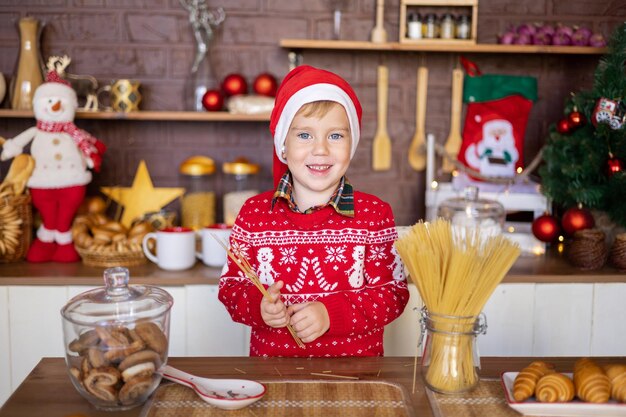 Een vrolijke baby met een kerstmuts en een rode trui viert Kerstmis in de keuken van het huis, prettige kerstdagen en gelukkig nieuwjaar