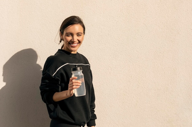 Een vrolijke blanke duizendjarige vrouw in sportkleding met een fles water die buiten rust, geniet van training