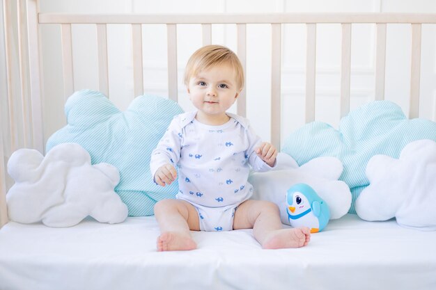 Een vrolijke blonde babyjongen in een wieg thuis met blauw en wit katoenen beddengoed glimlacht in de ochtend het concept van kinderartikelen en accessoires