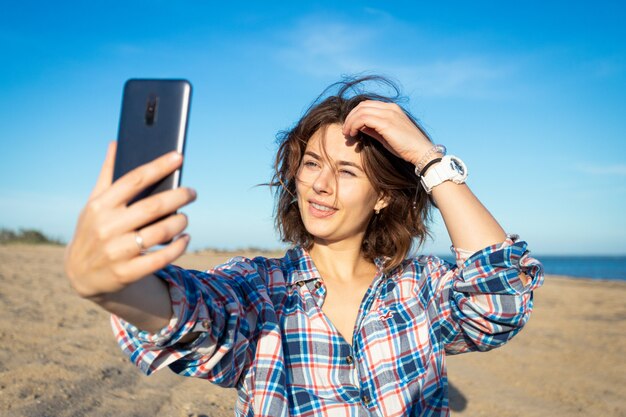 Een vrolijke donkerharige vrouw lacht, maakt een selfie aan de telefoon, loopt langs het strand, geniet van de felle zon op een zomerse dag. concept van zomervakantie op zee en live stijl