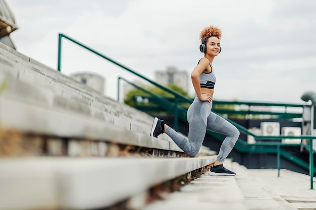 Een vrolijke gemotiveerde fitte sportvrouw met krullend haar die naar muziek luistert en lunges doet op de trap