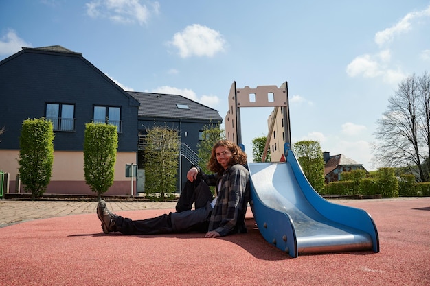 Een vrolijke jonge blanke man die bij de kinderglijbanen in de speeltuin zit