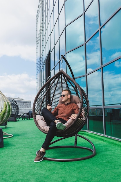 Een vrolijke jonge man in een shirt met een telefoon zit op het terras van een kantoorgebouw na het werk maakt gebruik van draadloos internet een positieve mannelijke werknemer met een bril praat aan de telefoon