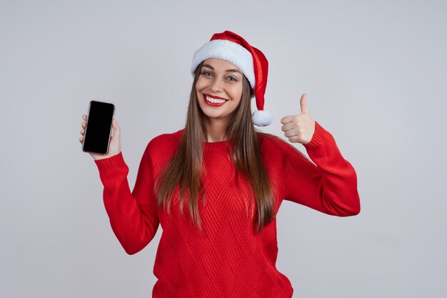 Een vrolijke jonge vrouw in een rode trui, met een kerstmuts op, laat haar duimen zien. de mobiele telefoon staat aan. Concept voor online felicitaties, verkoop, winkelen, kortingen.