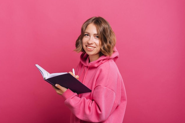 Een vrolijke jonge vrouw lacht naar de camera terwijl ze in haar planner schrijft in de buurt van een roze muur