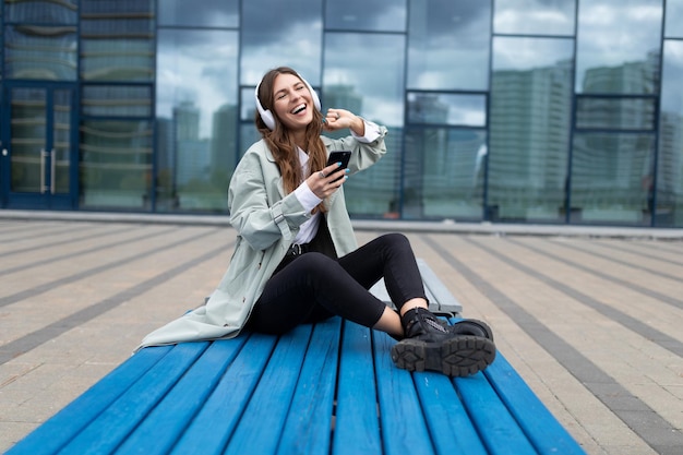 Een vrolijke jonge vrouw luistert naar muziek in een koptelefoon met een telefoon terwijl ze voor de