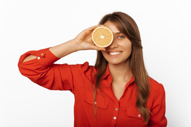 Een vrolijke jonge vrouw met een rood shirt bedekt een oog met een halve sinaasappel