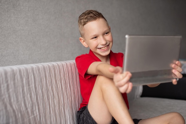 Een vrolijke jongen in een rood T-shirt zit op de bank met een tablet en lacht