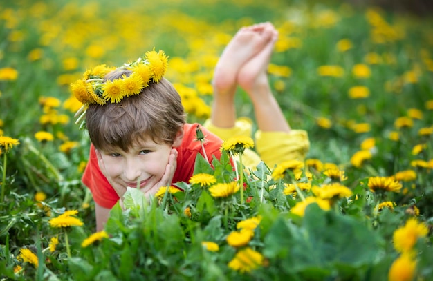 Een vrolijke jongen met een krans van paardebloemen ligt op het gras Kind in de wei