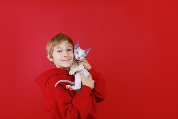 Een vrolijke jongen op een rode achtergrond houdt een klein wit katje vast, drukt hem tegen zich aan en knuffelt