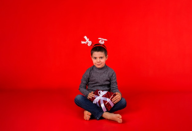 Een vrolijke jongen peuter in een trui en een kerst hoofdband met rendieren en met een cadeau zit op rood